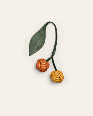 Yellow and orange leather bag charm in the shape of a cherry with a green leaf on a beige background