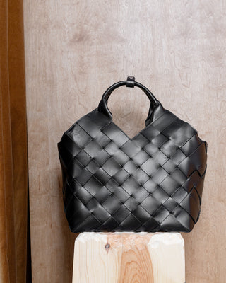 A black woven leather handbag with a structured basketweave pattern and a single top handle, displayed on a light wood block against a wood-paneled background.