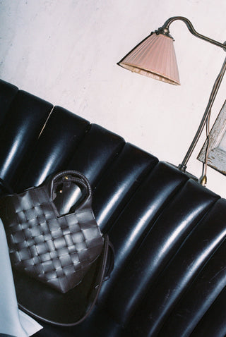 A dark woven leather handbag with a top handle rests on a black leather ribbed sofa in a dimly lit room, with a wall lamp visible in the background.
